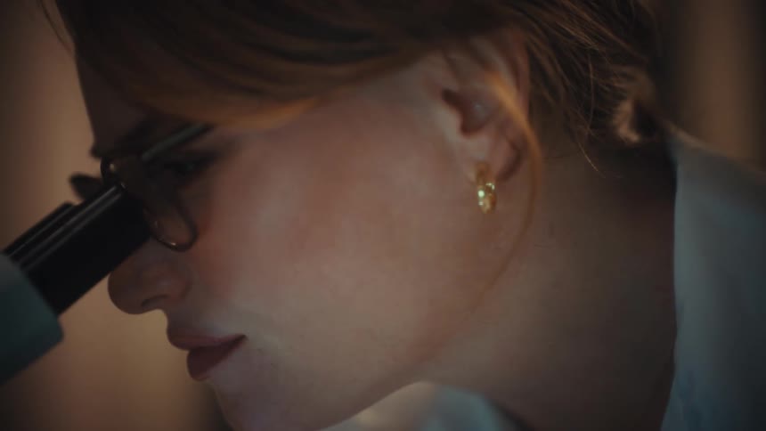 A close-up of a female scientist in glasses intently looking through a microscope in a dimly lit laboratory