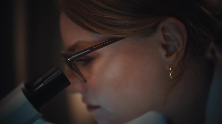 A focused female scientist with glasses carefully prepares and examines a slide under a microscope in a dark laboratory
