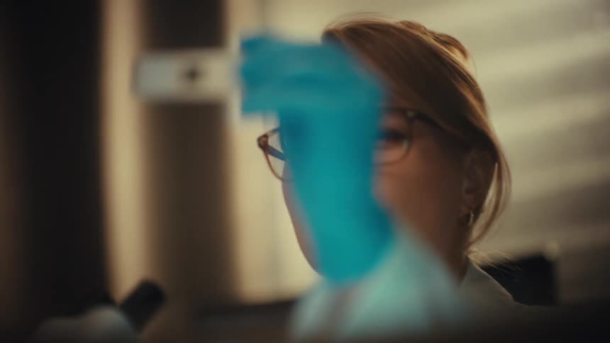 A focused female scientist with glasses carefully examines a sample on a glass slide in a dimly lit laboratory, ending with a slow zoom into a close-up of her face