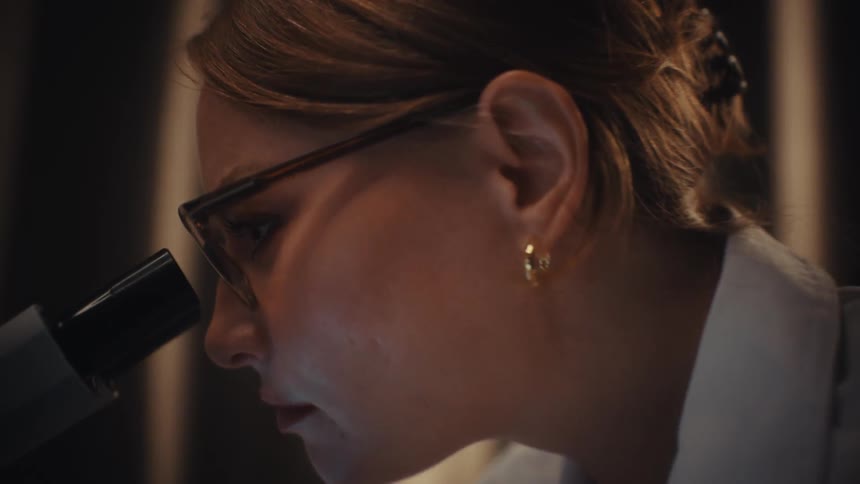 A focused female scientist in a lab coat meticulously examines a sample through a microscope in a dimly lit laboratory