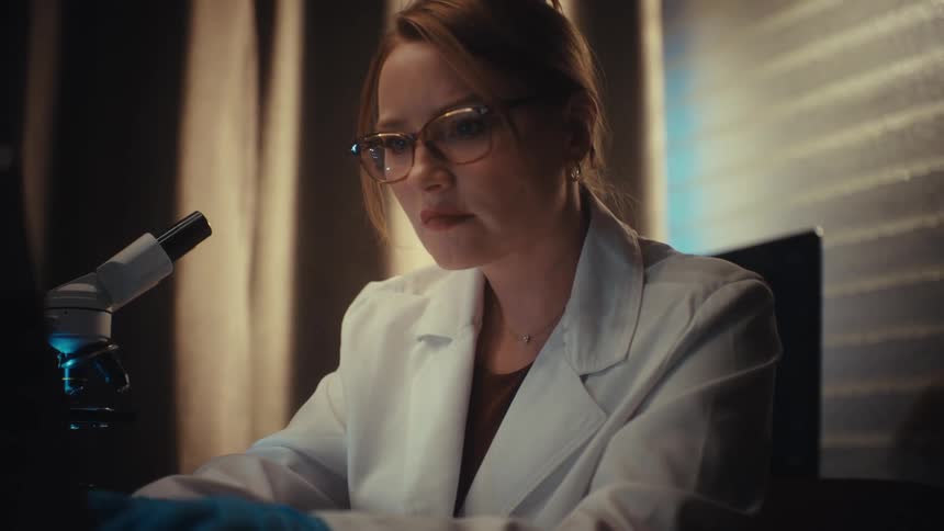A female scientist in a lab coat and glasses uses a microscope and then analyzes her findings on a computer screen in a dark laboratory