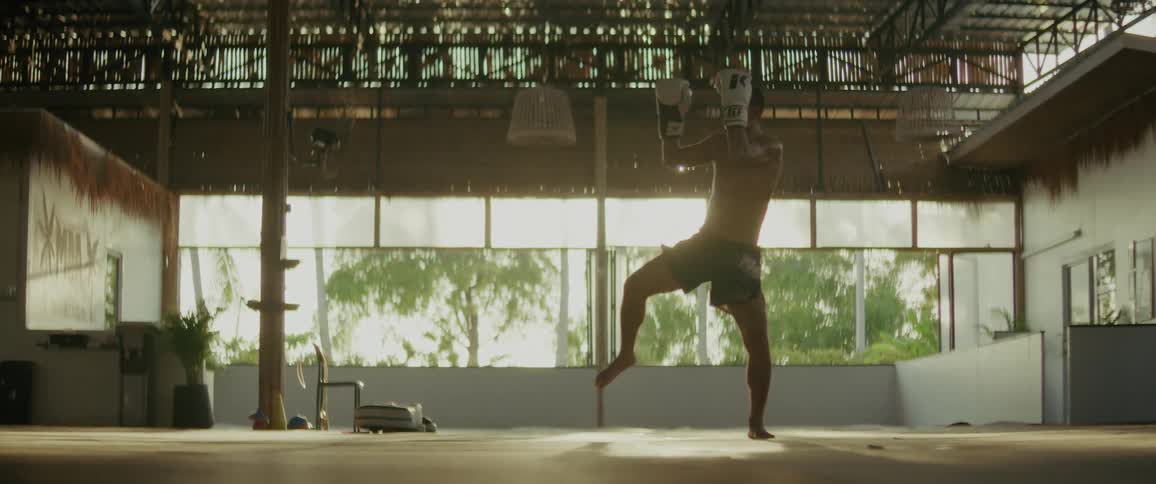 A shirtless Muay Thai fighter practices powerful kicks and punches in dramatic slow motion in a sunlit, rustic tropical gym