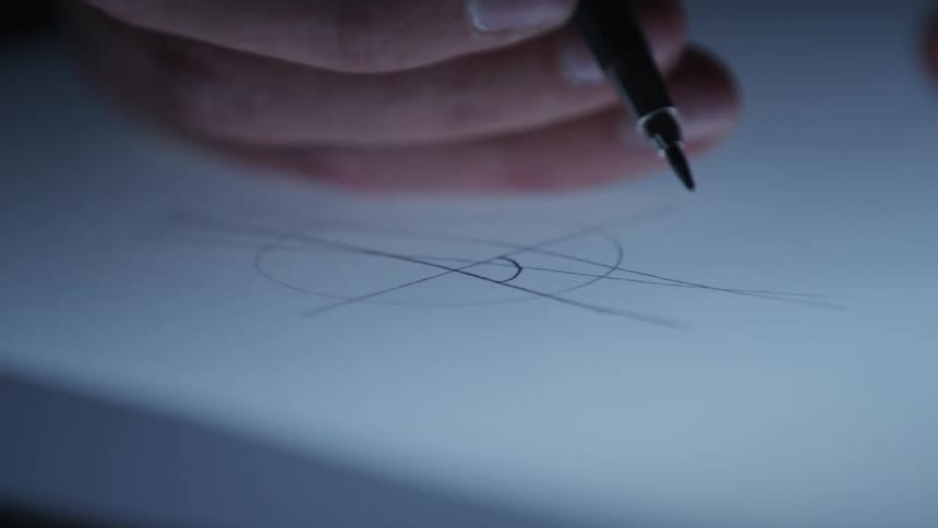 An architect's hand holding a fine tip pen hovers over a geometric drawing on a backlit table