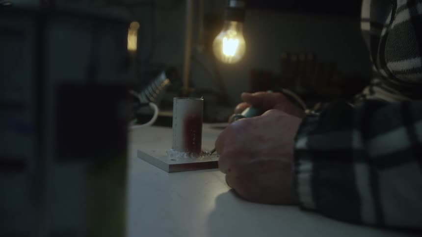 A person in a plaid shirt uses a rotary tool to meticulously work on a small project in a dimly lit workshop
