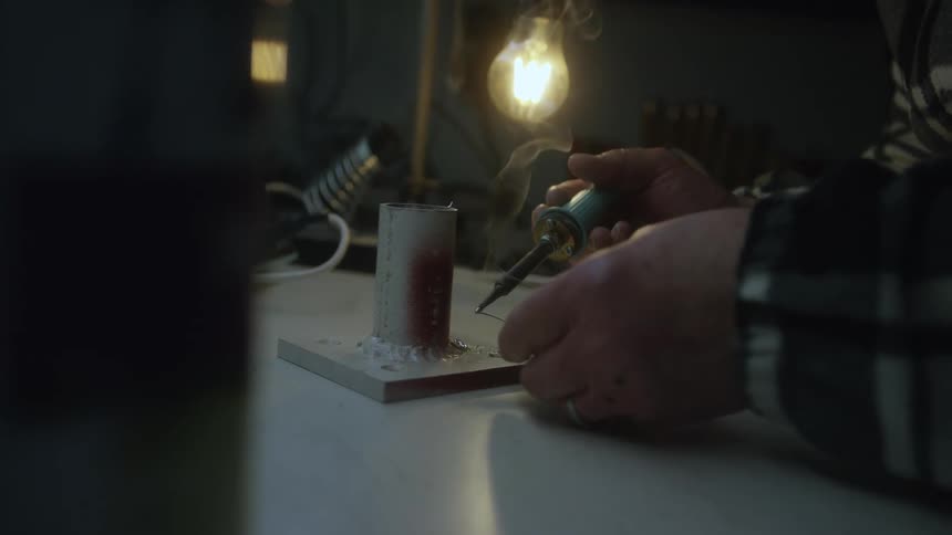 A craftsman carefully solders a metal piece on a workbench under the warm glow of a single lightbulb