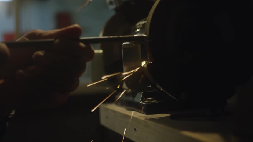 A close-up slow-motion shot of a metal rod being sharpened on a bench grinder, sending a cascade of bright orange sparks flying in a dark workshop