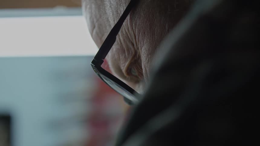 An extreme close-up side profile of an elderly man wearing glasses looking down in deep concentration