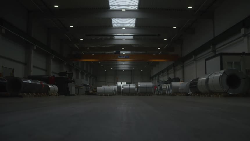 A slow upward tilt reveals a vast, dimly lit industrial warehouse filled with large rolls of sheet metal