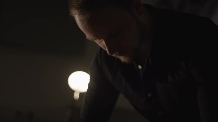 A serious man works diligently in a dimly lit room with only a small lamp for light