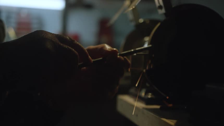 A person's hands carefully sharpen a metal tool on a grinding wheel in a dimly lit workshop, sending sparks flying in slow motion