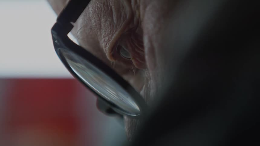 A dramatic extreme close-up reveals the focused, wrinkled eye of an elderly person wearing glasses