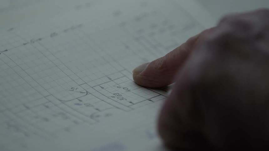 An engineer's finger follows the specifications on a detailed, hand-drawn mechanical blueprint