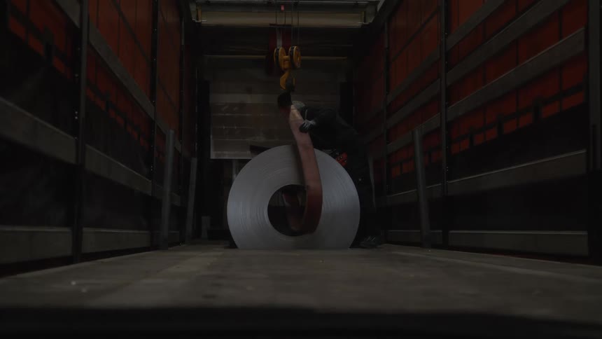 A factory worker uses an overhead crane to carefully load a large coil of sheet metal into the back of a truck
