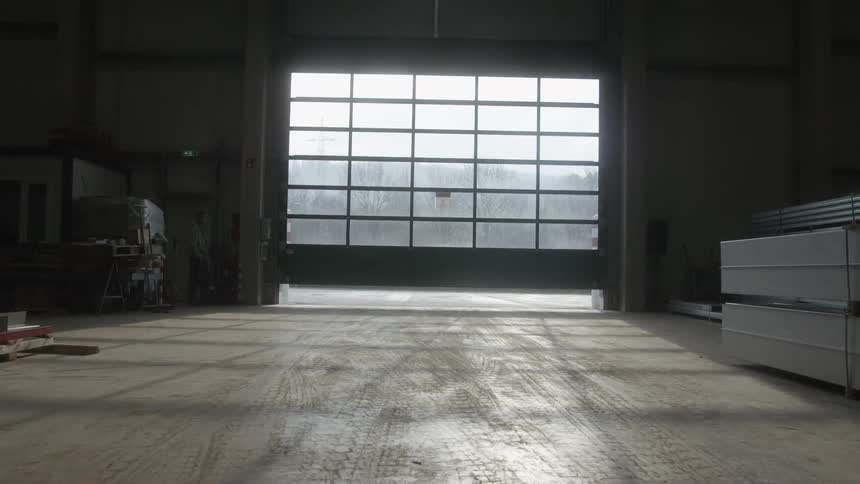 A large automatic rolling garage door closes on a warehouse, slowly darkening the empty interior