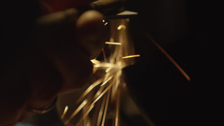 Sparks fly in extreme close-up slow motion as a metal tool is sharpened on a grinding wheel in a dark workshop