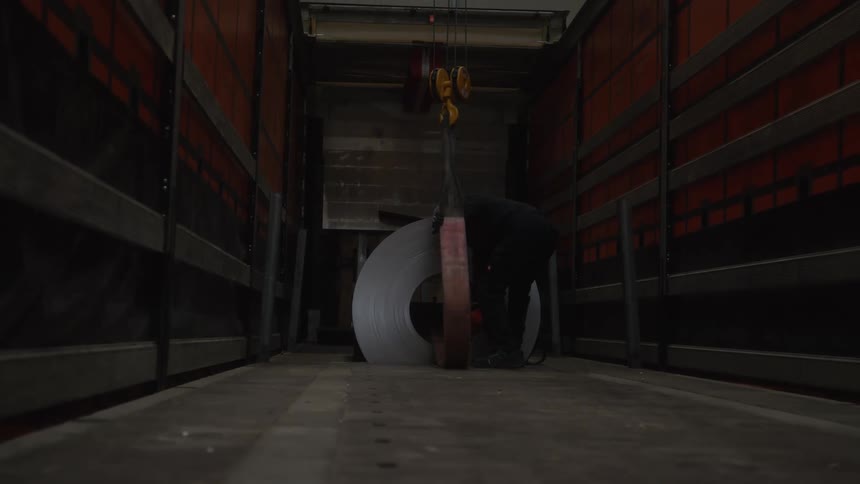A factory worker uses an overhead crane to carefully position a heavy roll of sheet steel inside the back of a transport truck