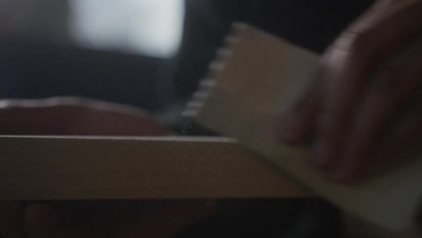 A close-up, slow-motion shot of a person sanding a wooden plank by hand, with sawdust flying through the air