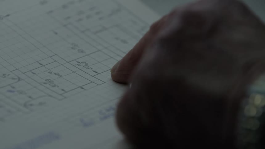 A close-up of a finger carefully tracing the dimensions on a hand-drawn technical blueprint on graph paper