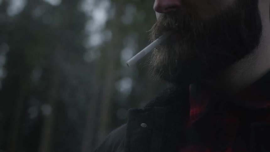 A bearded man in a flannel shirt exhales a thick cloud of cigarette smoke in slow motion