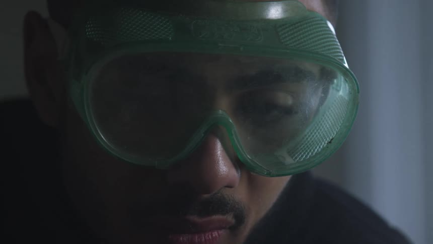 A close-up shot of a focused man wearing green safety goggles while concentrating on his work in a dimly lit setting