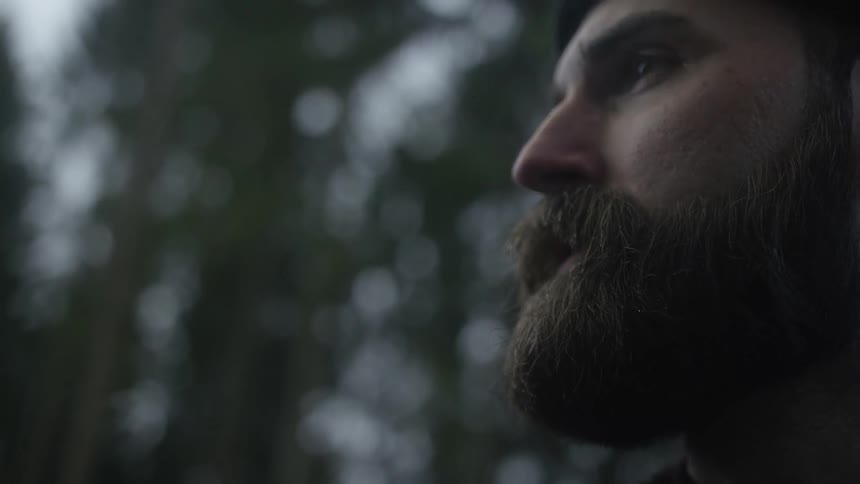 A bearded man takes a slow-motion drag of a cigarette and exhales a plume of smoke on a moody, overcast day