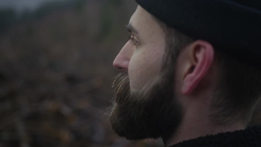 A slow-motion, shallow focus profile shot of a contemplative bearded man in a beanie looking into the distance