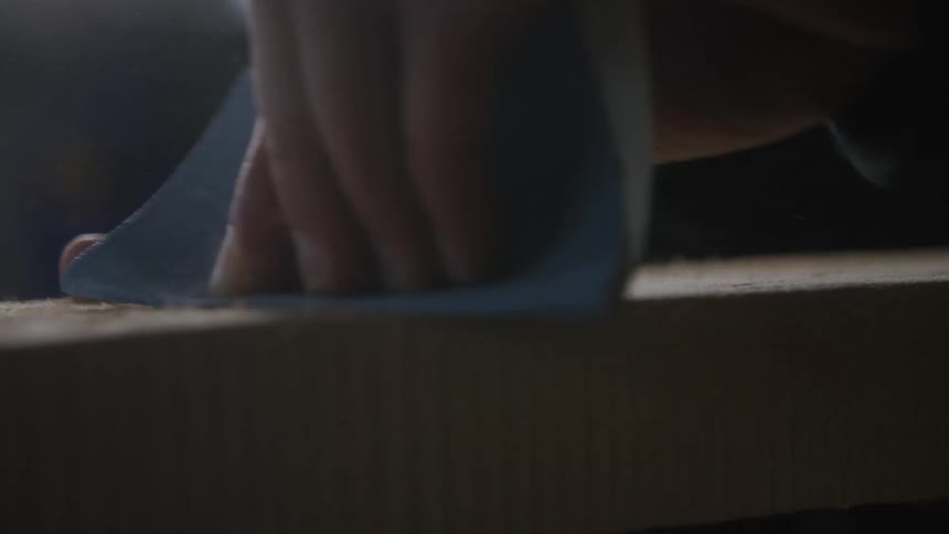 A close-up slow motion shot of a craftsman's hand sanding a wooden plank, creating a cloud of fine sawdust