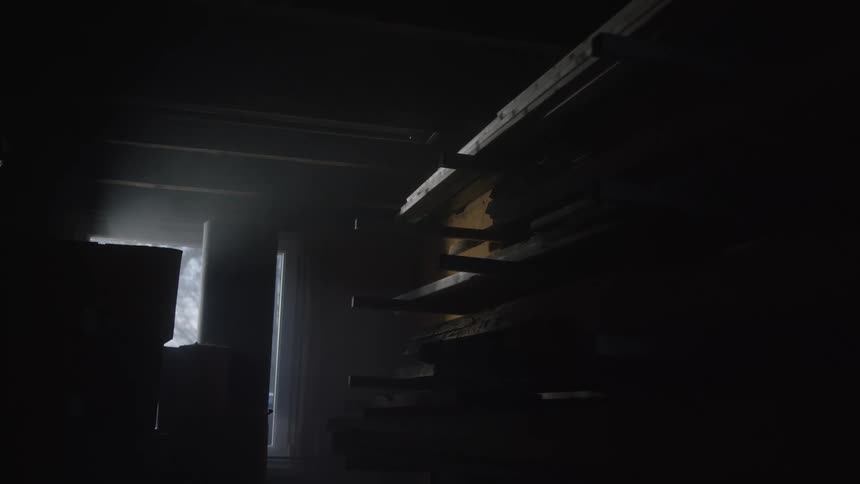Eerie god rays of light shine through a window into a dark, dusty woodworking shop with stacked lumber