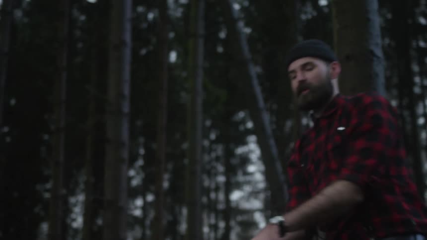 A bearded lumberjack in a red plaid shirt forcefully swings his axe, chopping into a fallen tree trunk in a dense forest
