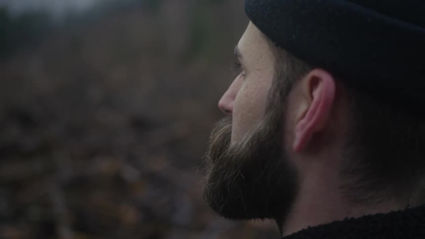 A close-up profile shot of a pensive bearded man wearing a black beanie, looking out thoughtfully in a somber, natural environment