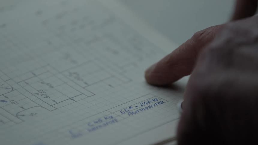 An engineer's finger slowly traces the lines of a hand-drawn technical blueprint on graph paper