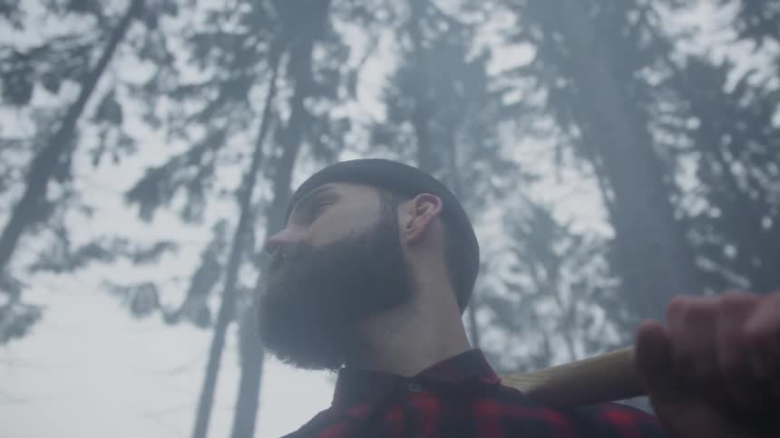 A bearded lumberjack with an axe on his shoulder looks up at the tall trees in a foggy, atmospheric forest