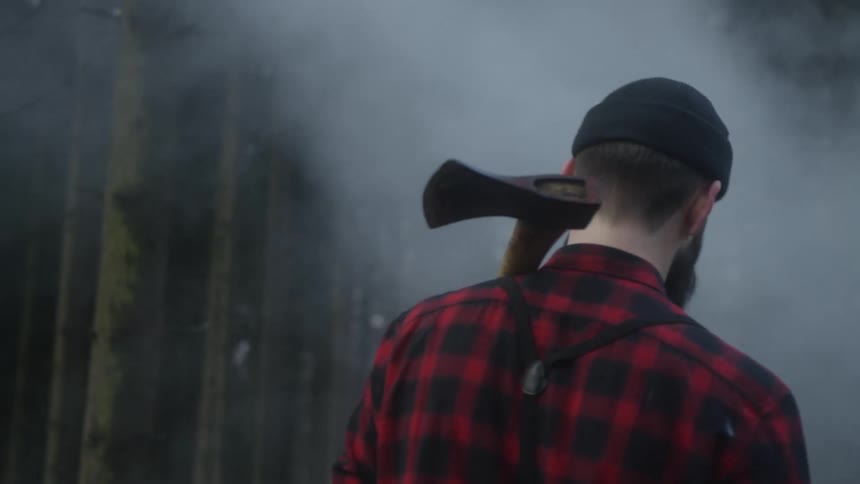 A bearded lumberjack in a plaid shirt walks slowly through a mysterious foggy forest with an axe over his shoulder