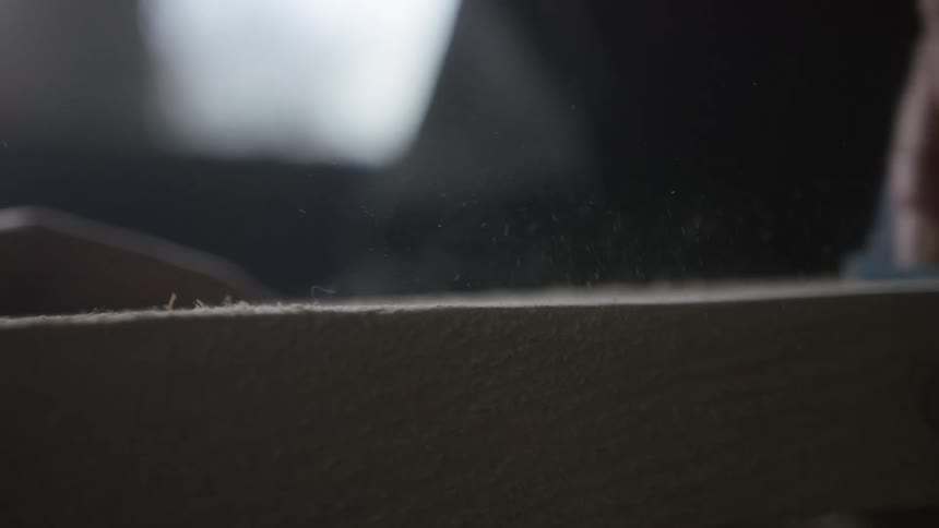 A close-up slow-motion shot shows a carpenter sanding a wooden plank, with dust flying into the air