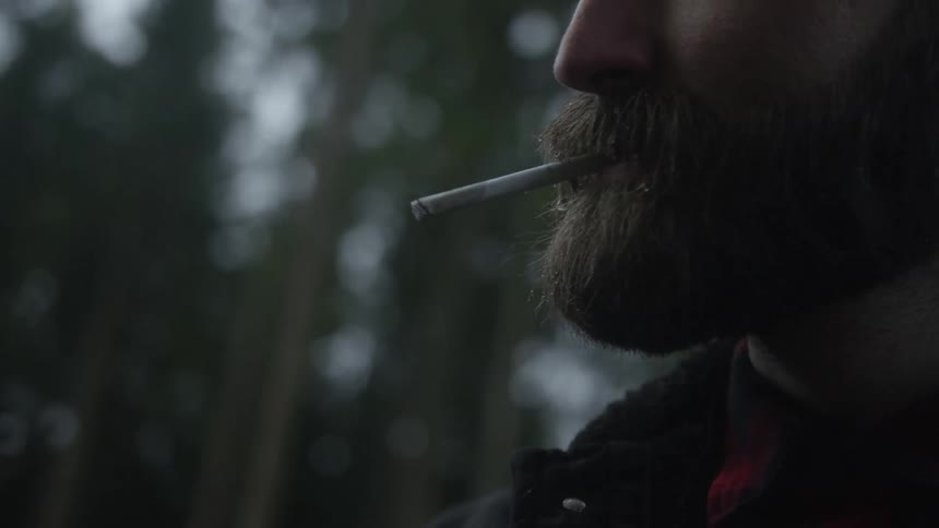 Close up side profile of a bearded man lighting a cigarette and exhaling smoke in a slow-motion outdoor shot