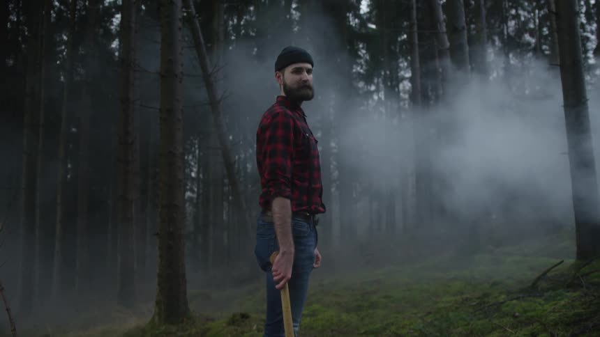 A bearded lumberjack stands with his axe amidst the swirling fog of a dark and mysterious forest