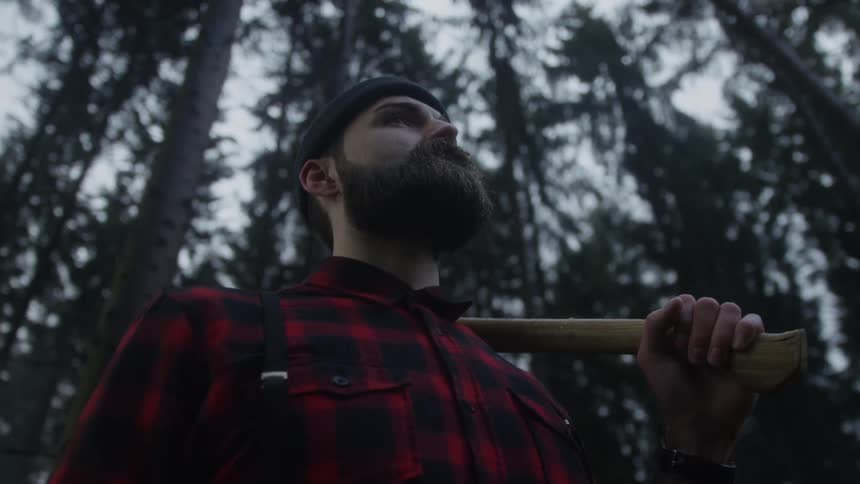 Low-angle dolly shot of a determined lumberjack with a beard holding an axe over his shoulder in a dark forest