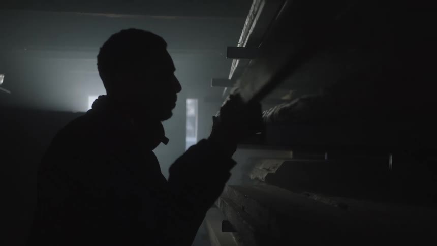 A silhouetted man in a dark, dusty workshop carefully selects and pulls a wooden plank from a shelf in slow motion