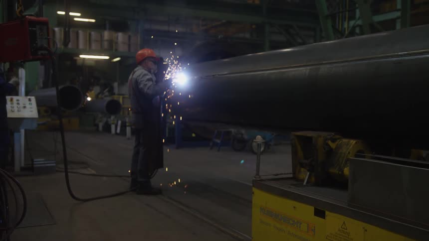 A welder in full protective gear works on a large metal pipe inside a dimly lit factory, with bright sparks illuminating the scene
