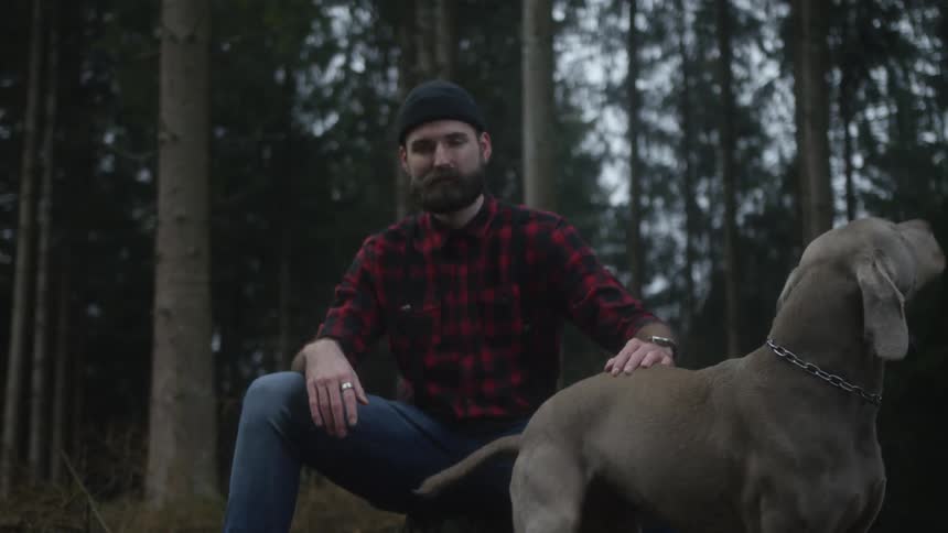 A bearded man in a plaid shirt sits in a dark forest while petting his Weimaraner dog
