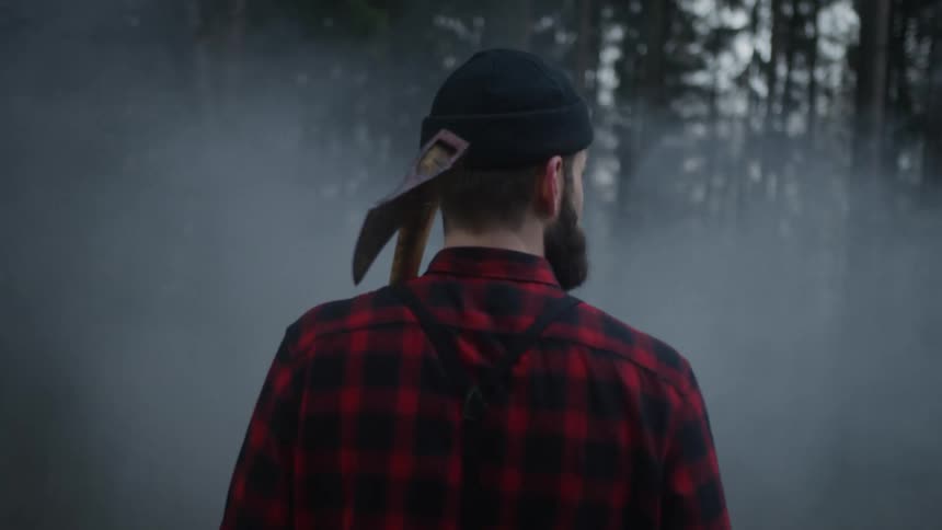 Back view of a rugged lumberjack carrying an axe over his shoulder as he ventures into a dense, smoky woodland