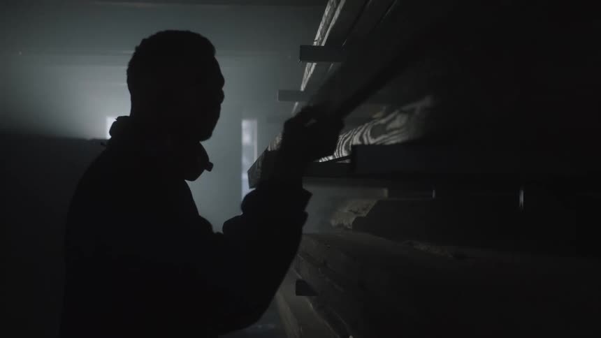 A silhouetted woodworker pulls a long wooden plank from a dusty shelf in a dark, atmospheric workshop