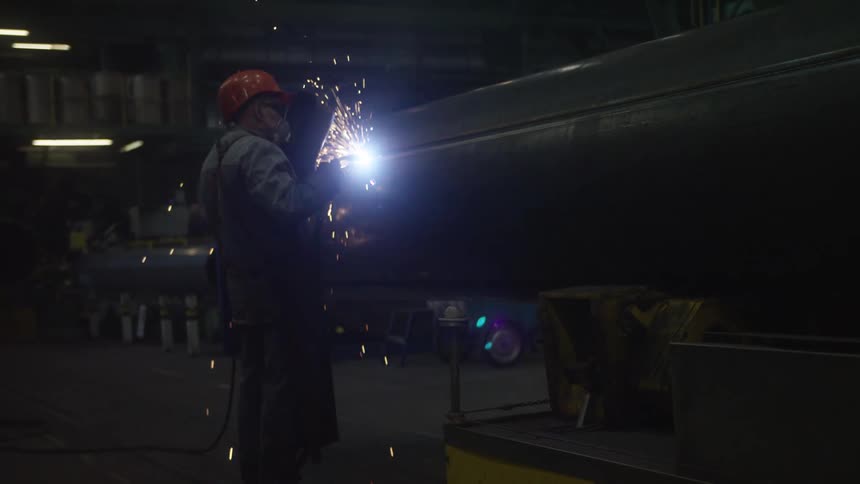 Slow motion footage shows a welder in a dark factory creating a shower of sparks as they work on a large metal pipe