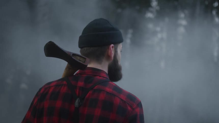 A lumberjack in a plaid shirt and beanie walks through a mysterious foggy forest with an axe over his shoulder in slow motion