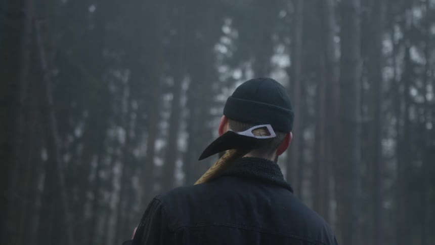 A mysterious man with an axe over his shoulder walks slowly into the depths of a dark, foggy forest