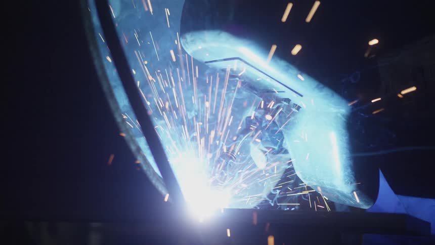 A welder in a protective mask works with a welding torch, creating bright blue light and golden sparks in a dark workshop