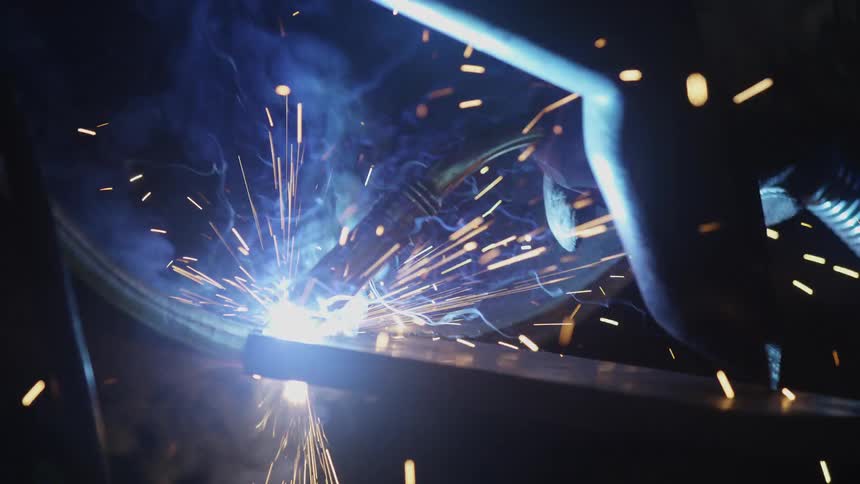 A close-up slow-motion shot of industrial welding shows brilliant sparks and smoke flying from the bright blue arc