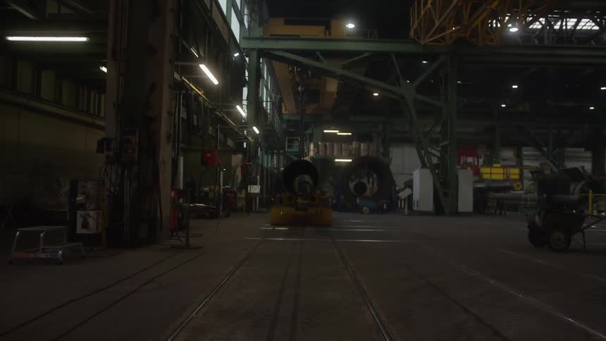 Sparks fly from automated machinery working on a large metal cylinder inside a dark, industrial factory