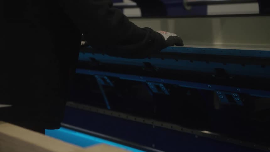 A factory worker wearing gloves carefully places a large sheet of metal into an industrial bending machine