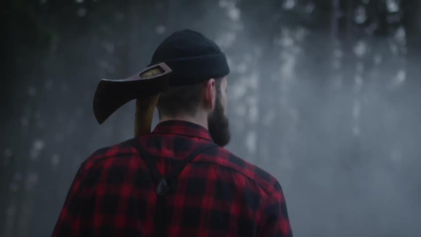 A lumberjack with a beard and an axe over his shoulder stands in a dark, foggy forest
